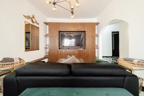 A modern living room with a large flat-screen TV, a wooden accent wall, a black leather sofa, and a green ottoman.