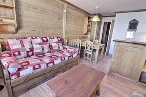 A cozy living area with a wooden sofa, a wooden coffee table, and a dining table with chairs. A kitchen area is visible in the background.