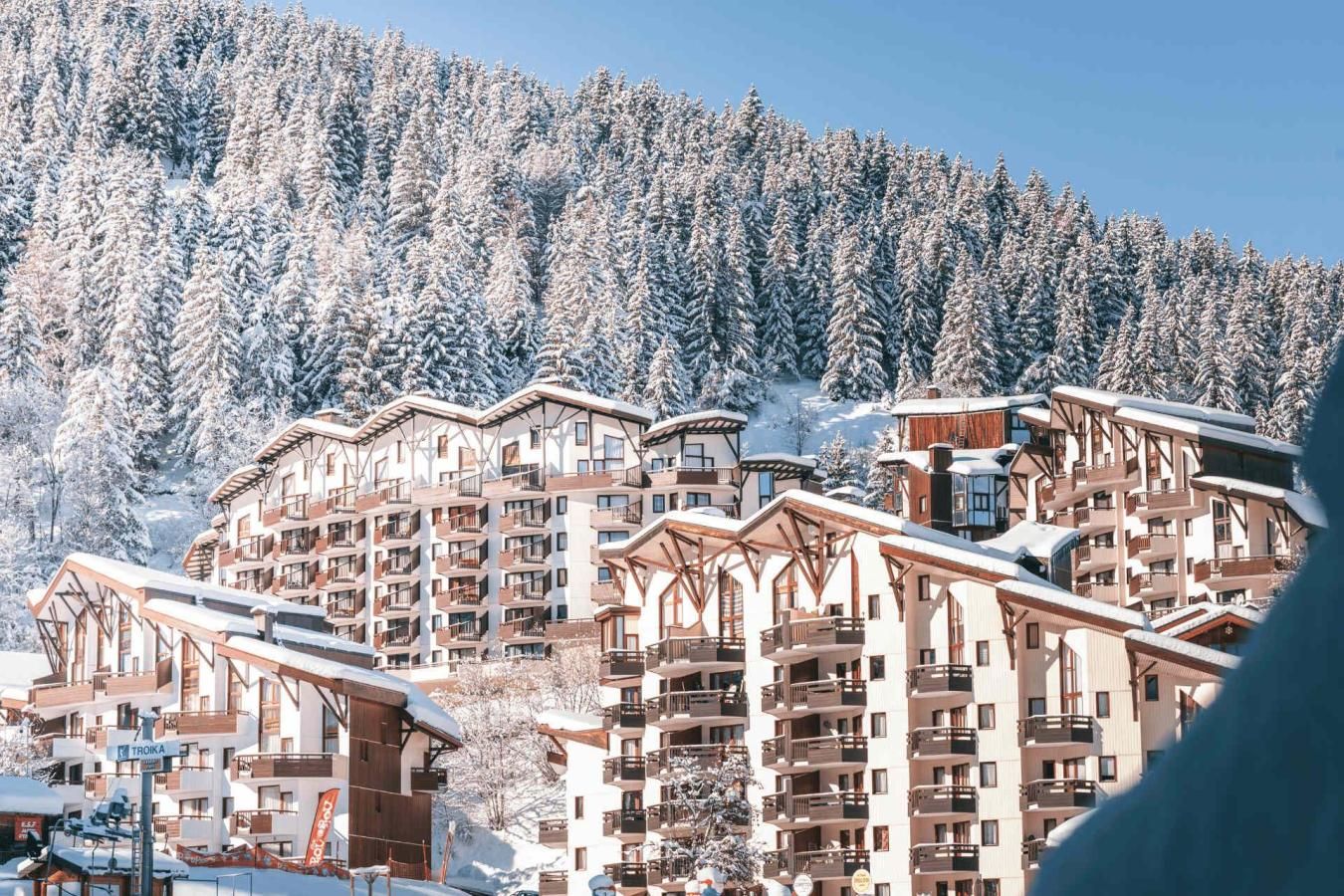 Une station de montagne enneigée, avec des bâtiments à plusieurs étages dotés de balcons, et des pistes de ski en arrière-plan.