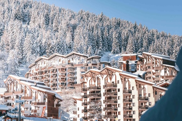 A snow-covered mountain resort with multi-story buildings featuring balconies and ski slopes in the background.