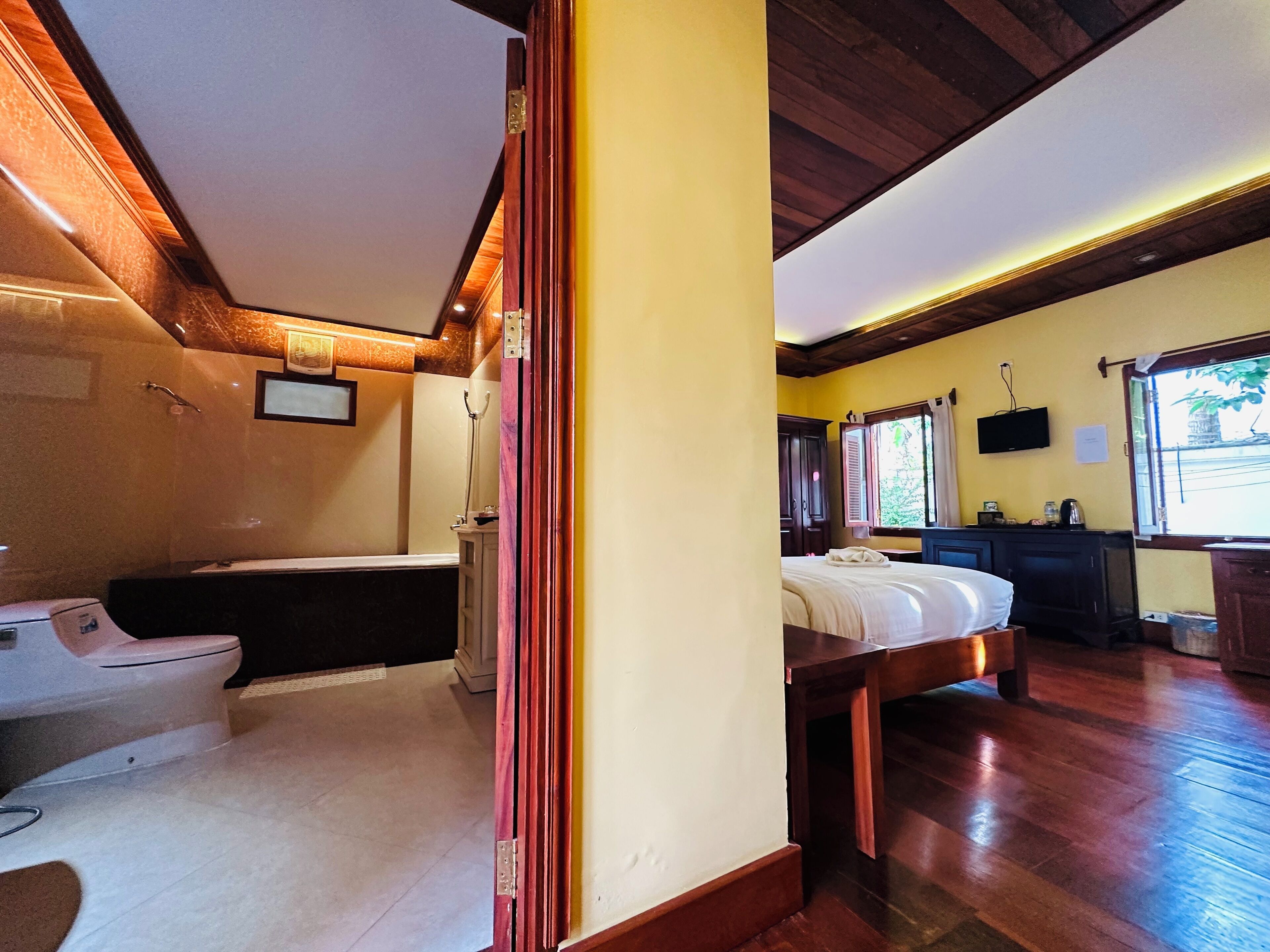 Quarto de hotel com piso de madeira, uma cama, escrivaninha, cadeira, janela e banheiro com vaso sanitário e pia.