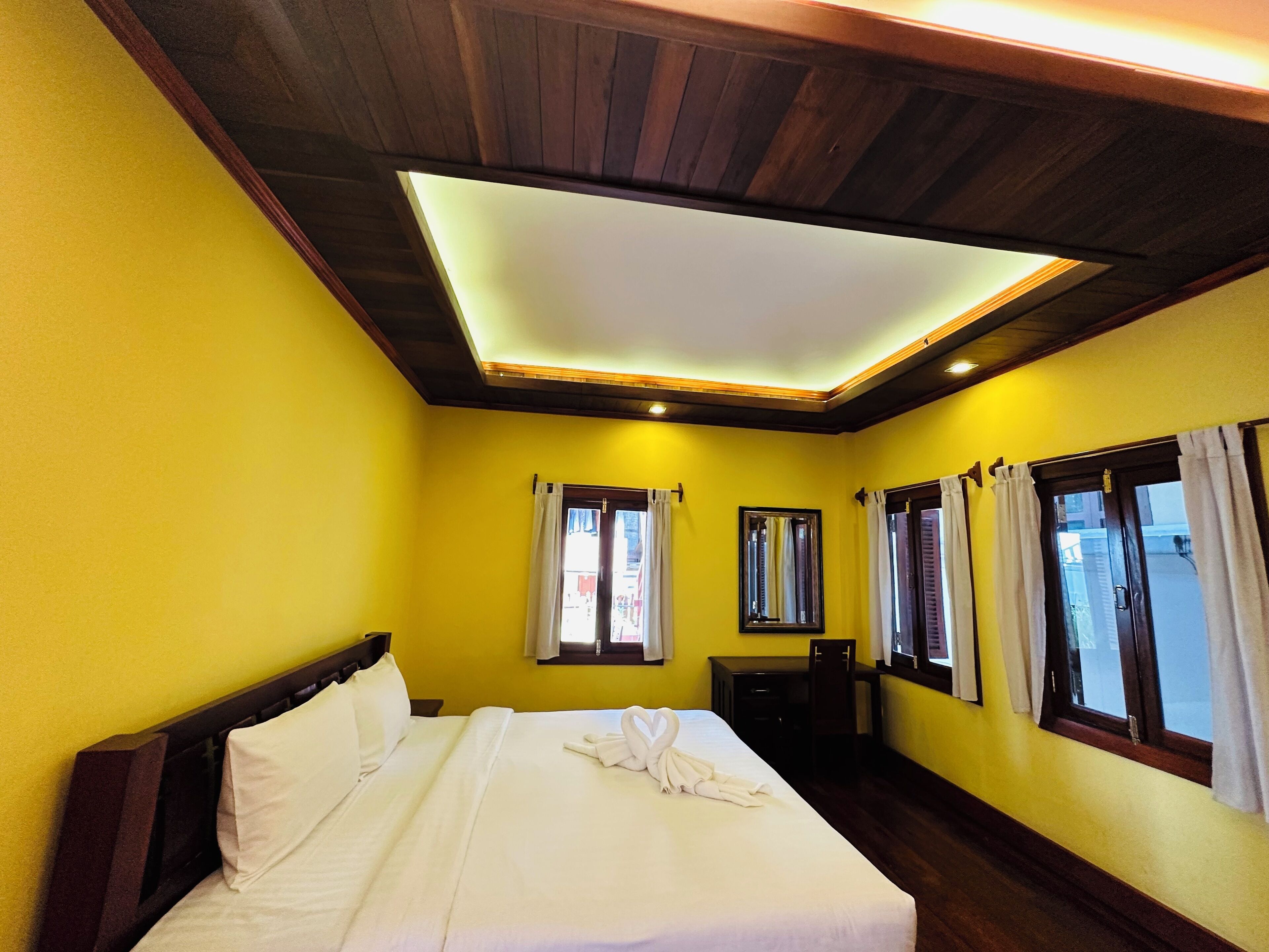 A hotel room with a wooden ceiling, yellow walls, a bed with white linens, a desk, and two windows with curtains.
