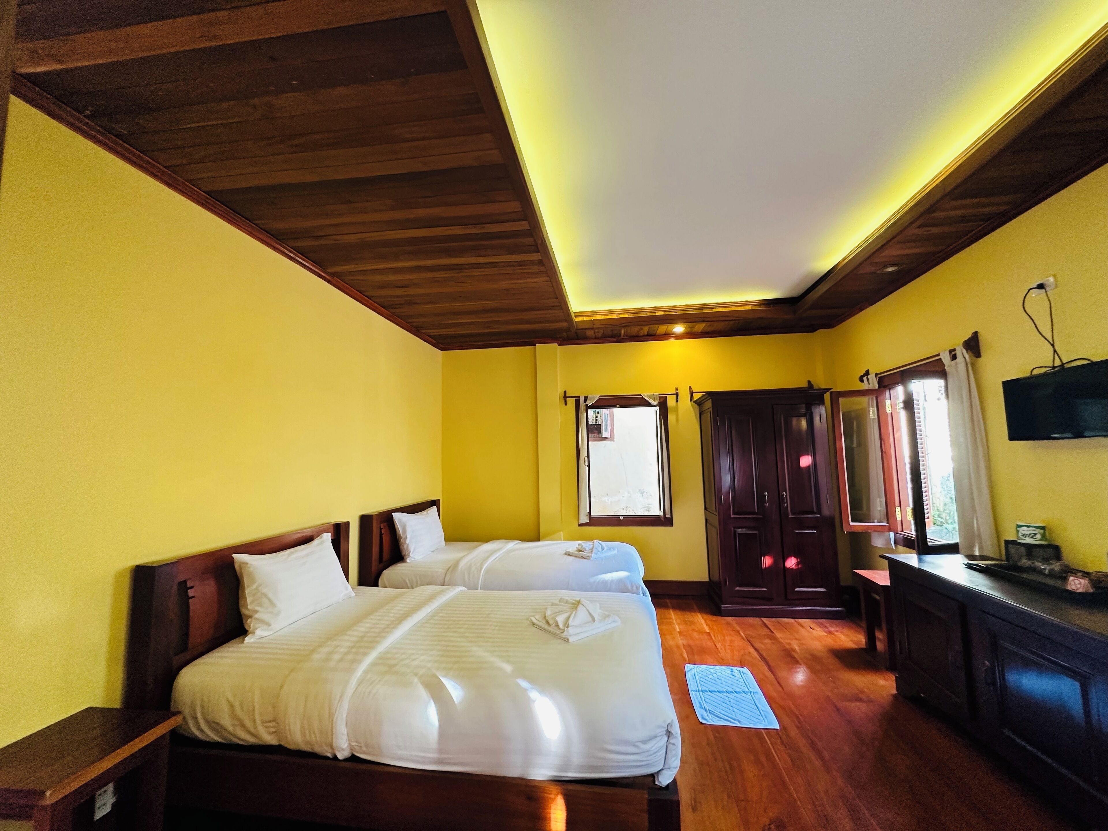 A hotel room with two beds, wooden ceiling, yellow walls, a window, a TV, and a wooden cabinet.