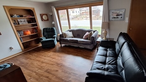 Een woonkamer met een houten vloer, een grijze bank, een zwarte leren zithoek, een boekenkast en een raam met uitzicht.