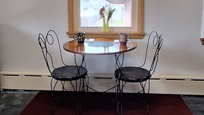 Een eethoek met een ronde tafel en twee stoelen, een raam met uitzicht op een huis, een klok aan de muur en een schilderij boven het raam.