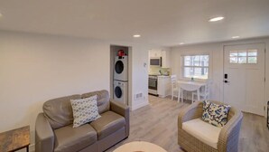 A compact living space with a sofa, a wicker chair, a kitchen area with a dining table, and a laundry machine. - 1Nt. Ok, Walk to Beach and Town, Romantic, Firepit, BBQ (Bungalow3) (Ocean Park)