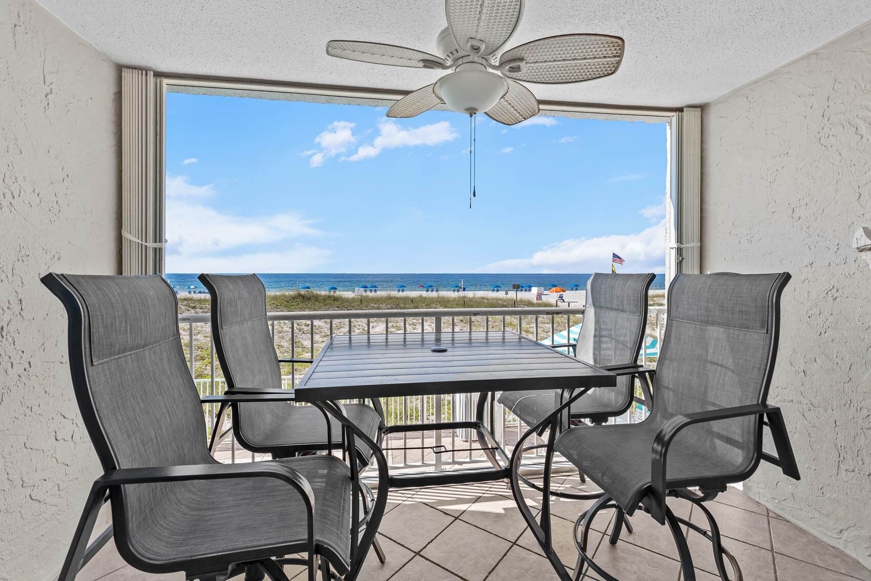 A balcony with a table and chairs, overlooking a beach with a clear sky and ocean view.
