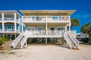 Une maison à deux étages située en bord de mer, dotée d’un balcon à l’étage supérieur, d’une terrasse couverte et d’un escalier menant à l’entrée.