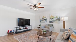 A modern living room with a flat-screen TV, a ceiling fan, a sofa, a coffee table, and two paintings on the wall. - Shared Pool- Beach Bungalow 2 (Fort Myers Beach)