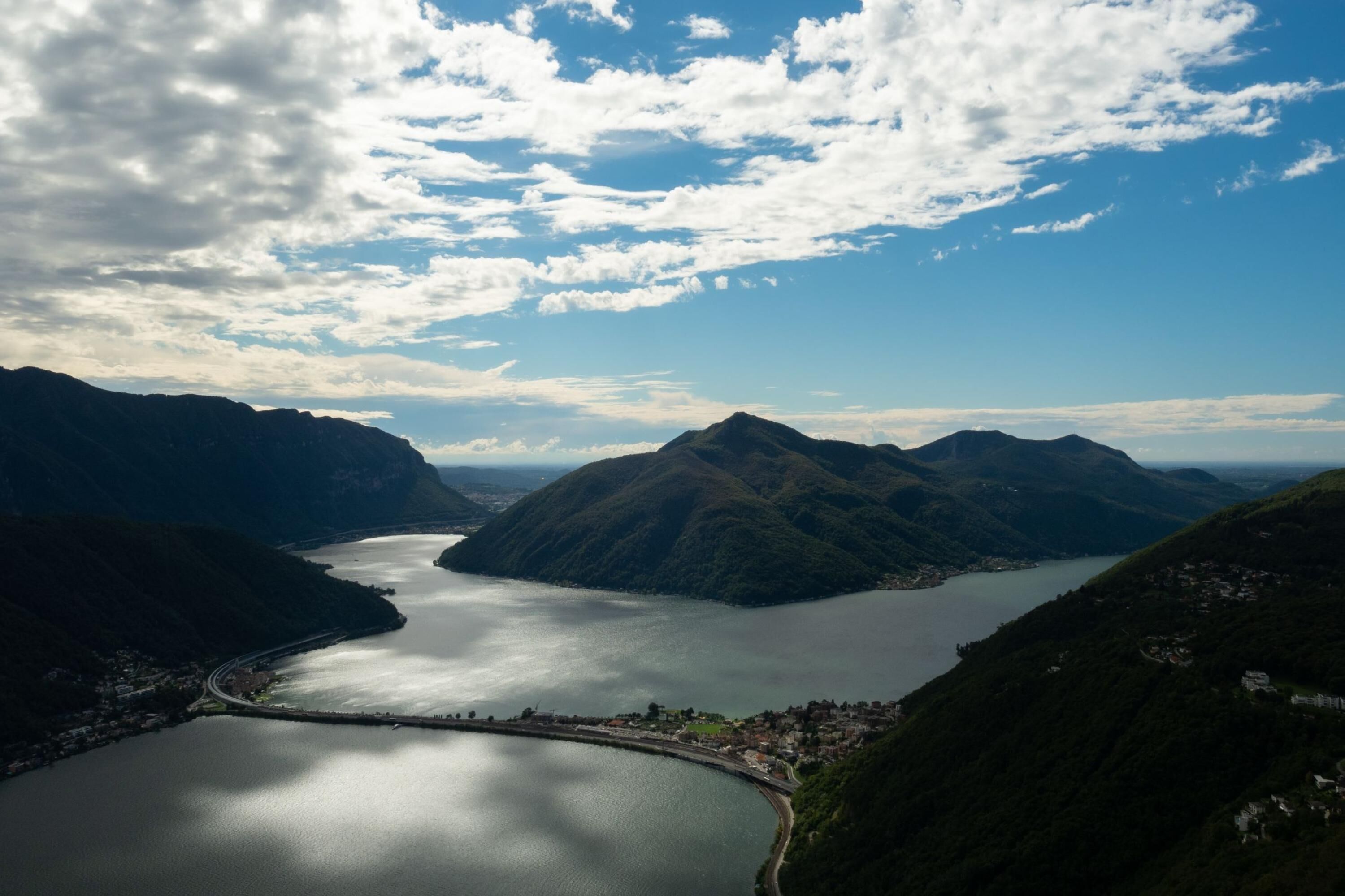 A panoramic view of a mountainous landscape with a large lake, a winding road, and a small town nestled among the hills.