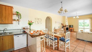 A kitchen with a dishwasher, wooden cabinets, a dining area with chairs, a sofa, and a TV.