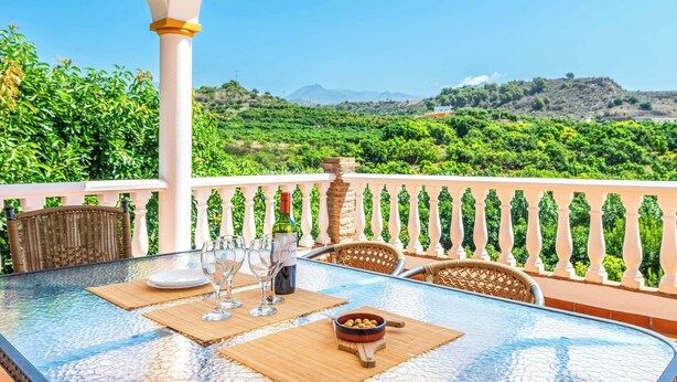 A table set with wine glasses and a bowl of olives on a balcony with a white railing and a view of a lush landscape.