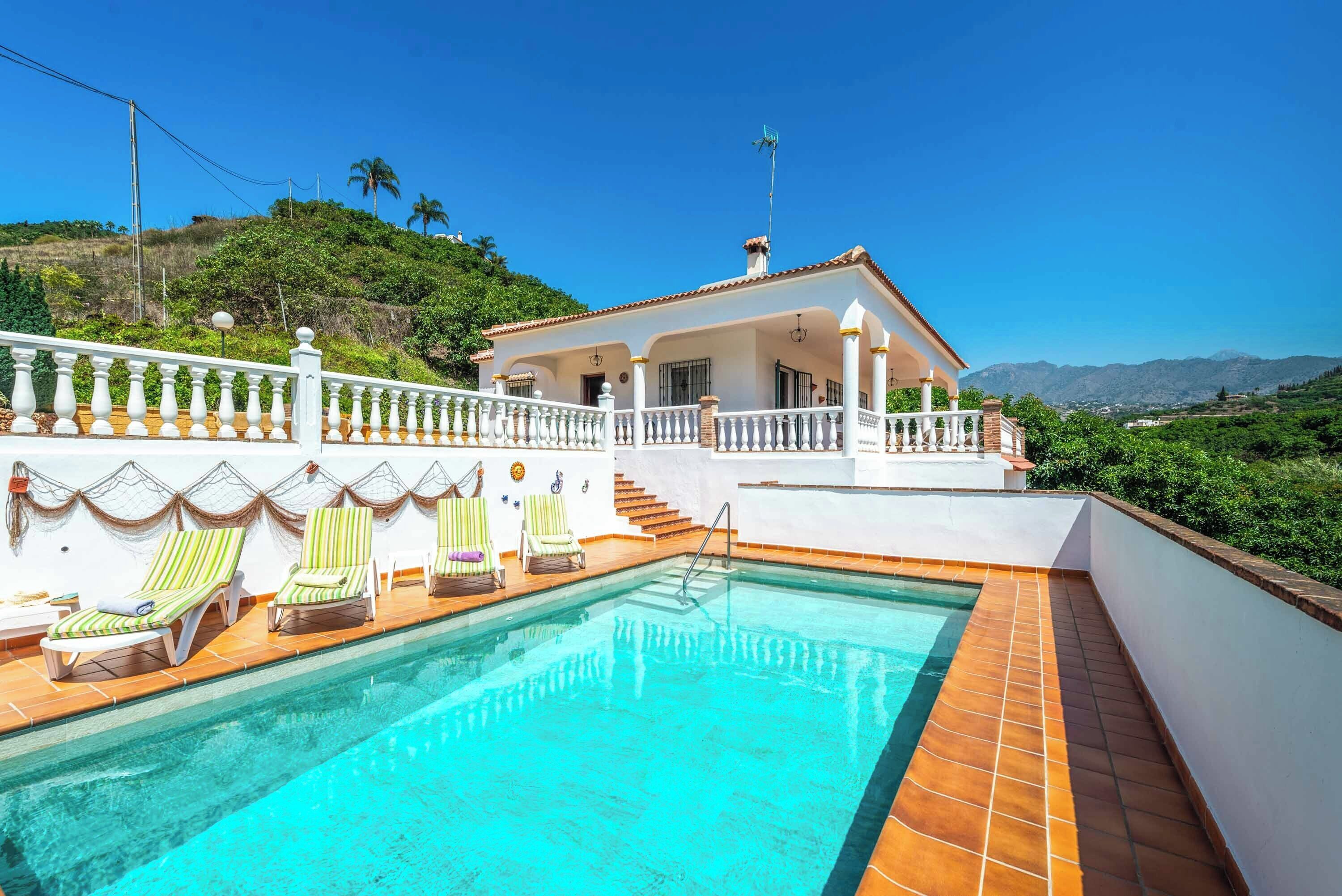 A pool area with lounge chairs, a white building with a red roof, and a view of hills and mountains.