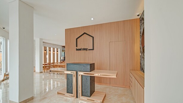 A modern hotel lobby with a reception desk, a wooden wall with a logo, and a seating area with tables and chairs. - Collection O 90774 Homi Stay (Surakarta)