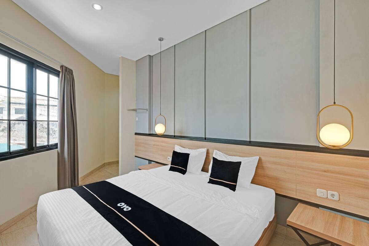 A modern hotel room with a large bed, bedside tables, a view through a window, and a wall-mounted light fixture.