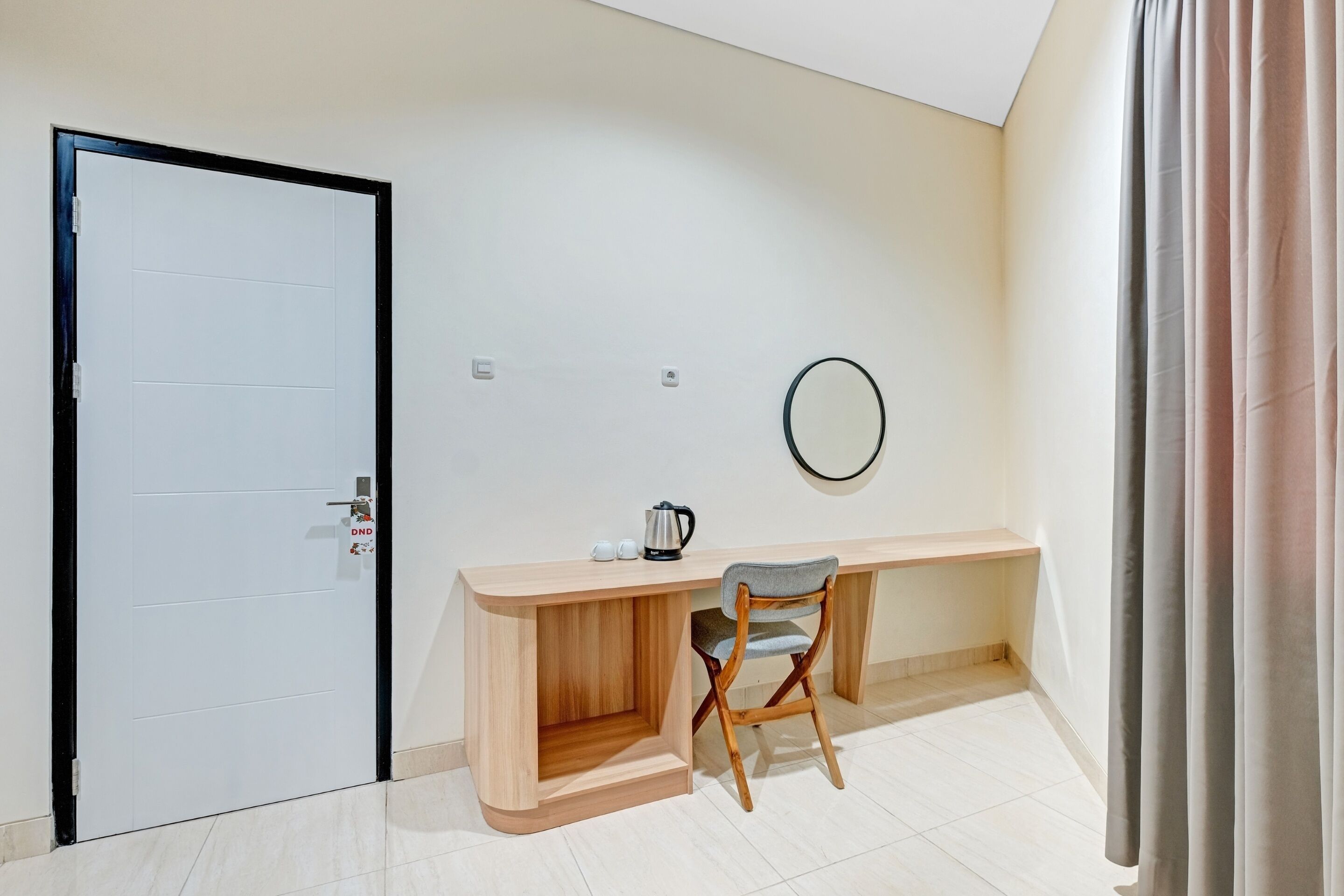 A room with a white door, a wooden desk with a chair, a small shelf, a round mirror, and a curtain.
