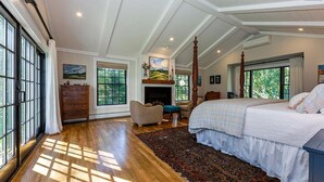 A spacious bedroom with a large bed, a fireplace, and a view of the outdoors through large windows. - Remodeled Beautiful 4-Bedroom Secluded Home in Downtown Bar Harbor! (Bar Harbor)