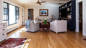 A spacious living room with a wooden floor, a ceiling fan, a large bookshelf, and a view through a window. - Remodeled Beautiful 4-Bedroom Secluded Home in Downtown Bar Harbor! (Bar Harbor)
