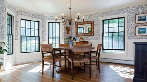 A dining room with a wooden table and chairs, large windows, and a chandelier. - Remodeled Beautiful 4-Bedroom Secluded Home in Downtown Bar Harbor! (Bar Harbor)