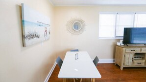 A room with a white table, chairs, a TV on a wooden cabinet, a painting on the wall, and a window with blinds.
