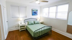 A bedroom with a single bed, two bedside tables, a ceiling fan, and large windows with blinds.