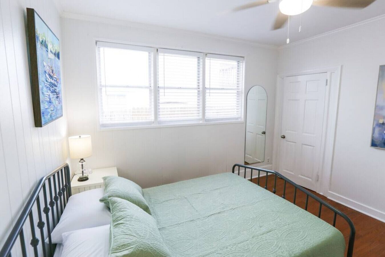 A bedroom with a single bed, a bedside table with a lamp, a window with blinds, a door, and a wall-mounted mirror.