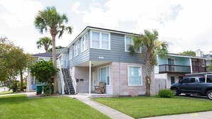 A two-story residential building with a covered front porch, a staircase leading to the upper level, and a vehicle parked in the driveway.