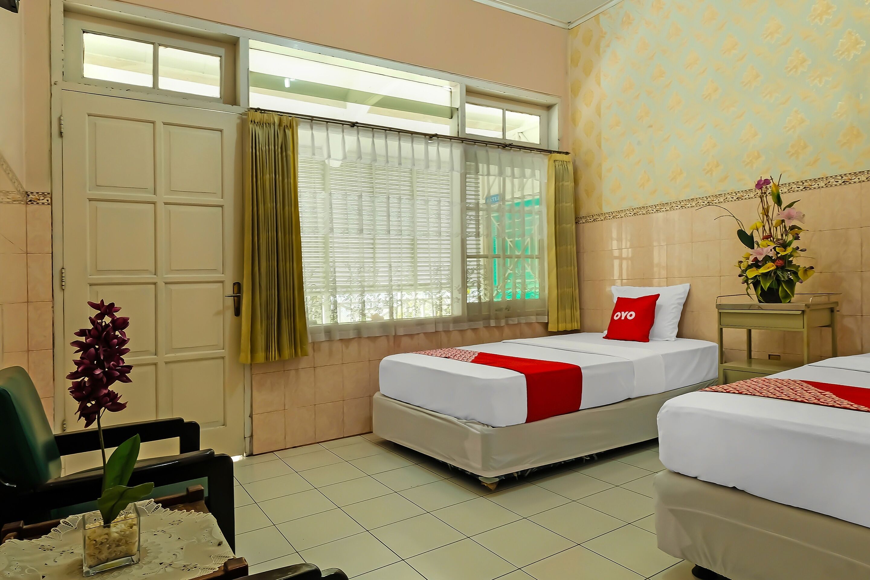 Habitación de hotel con dos camas, una ventana con cortinas, una mesita con un florero y una silla.