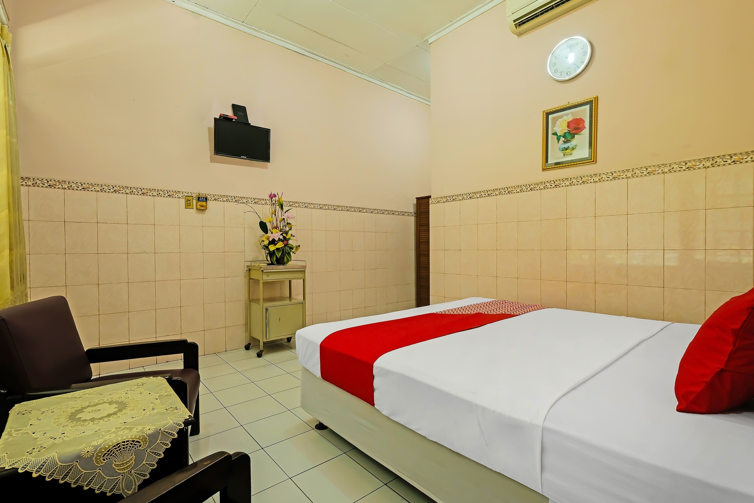 Ein Hotelzimmer mit Bett, Sessel, kleinem Tisch mit Spitze und einem wandmontierten Fernseher. Außerdem ist eine Uhr und ein gerahmtes Bild an der Wand zu sehen.