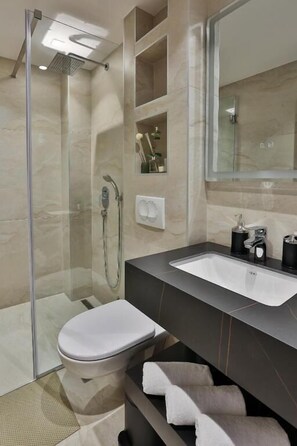 Un baño moderno con ducha envuelta en vidrio, inodoro blanco, lavamanos sobre encimera oscura y un espejo grande.