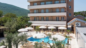 Un hotel de varios pisos con zona de piscina, mobiliario exterior y montañas al fondo.