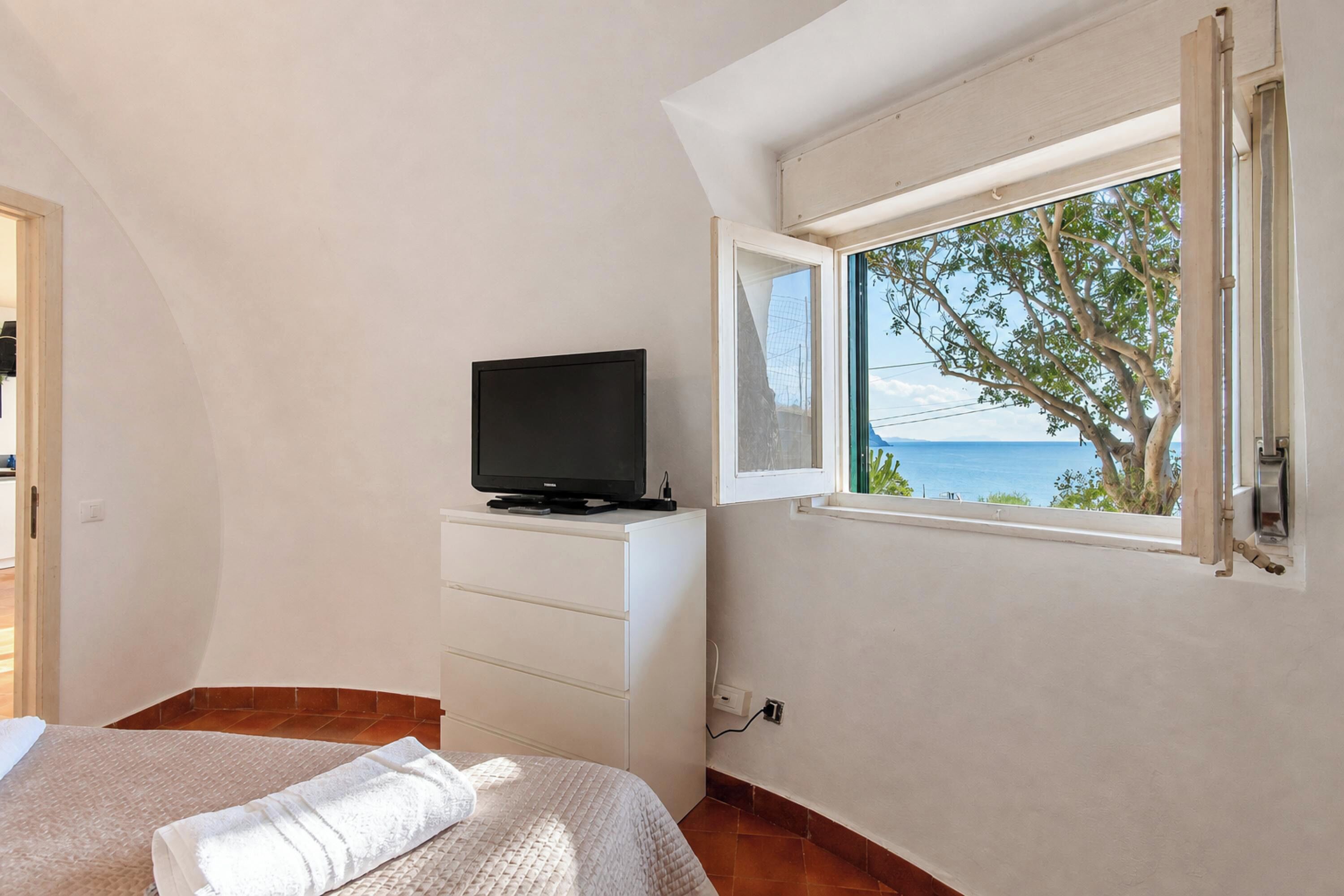 A bedroom with a bed, a TV on a dresser, a window with a view of the sea, and a door leading to another room.