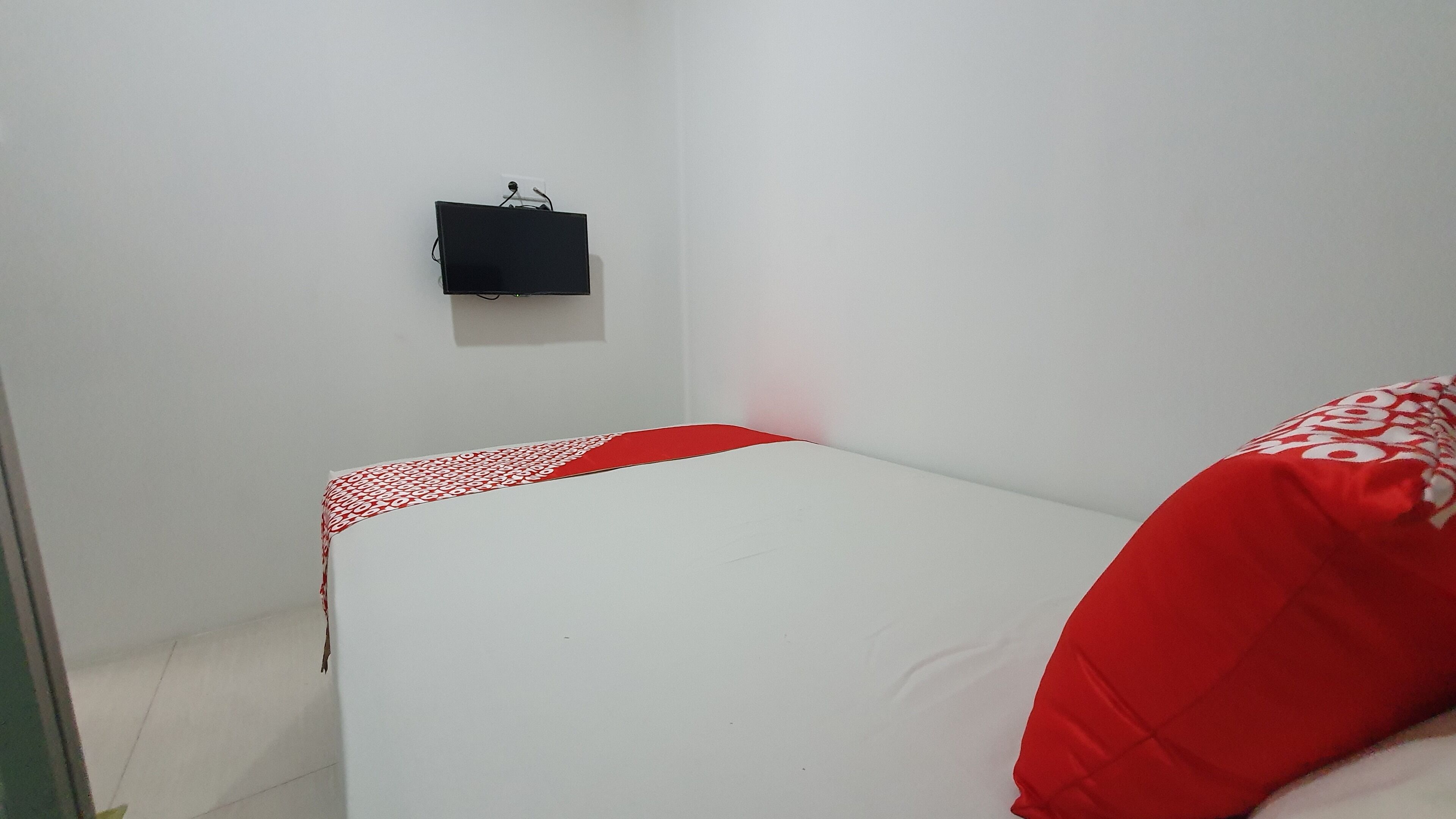 Una habitación pequeña con cama individual, colchón blanco y ropa de cama roja. Un altavoz negro está montado en la pared.