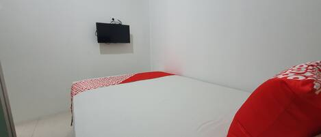 Una habitación pequeña con cama individual, colchón blanco y ropa de cama roja. Un altavoz negro está montado en la pared.