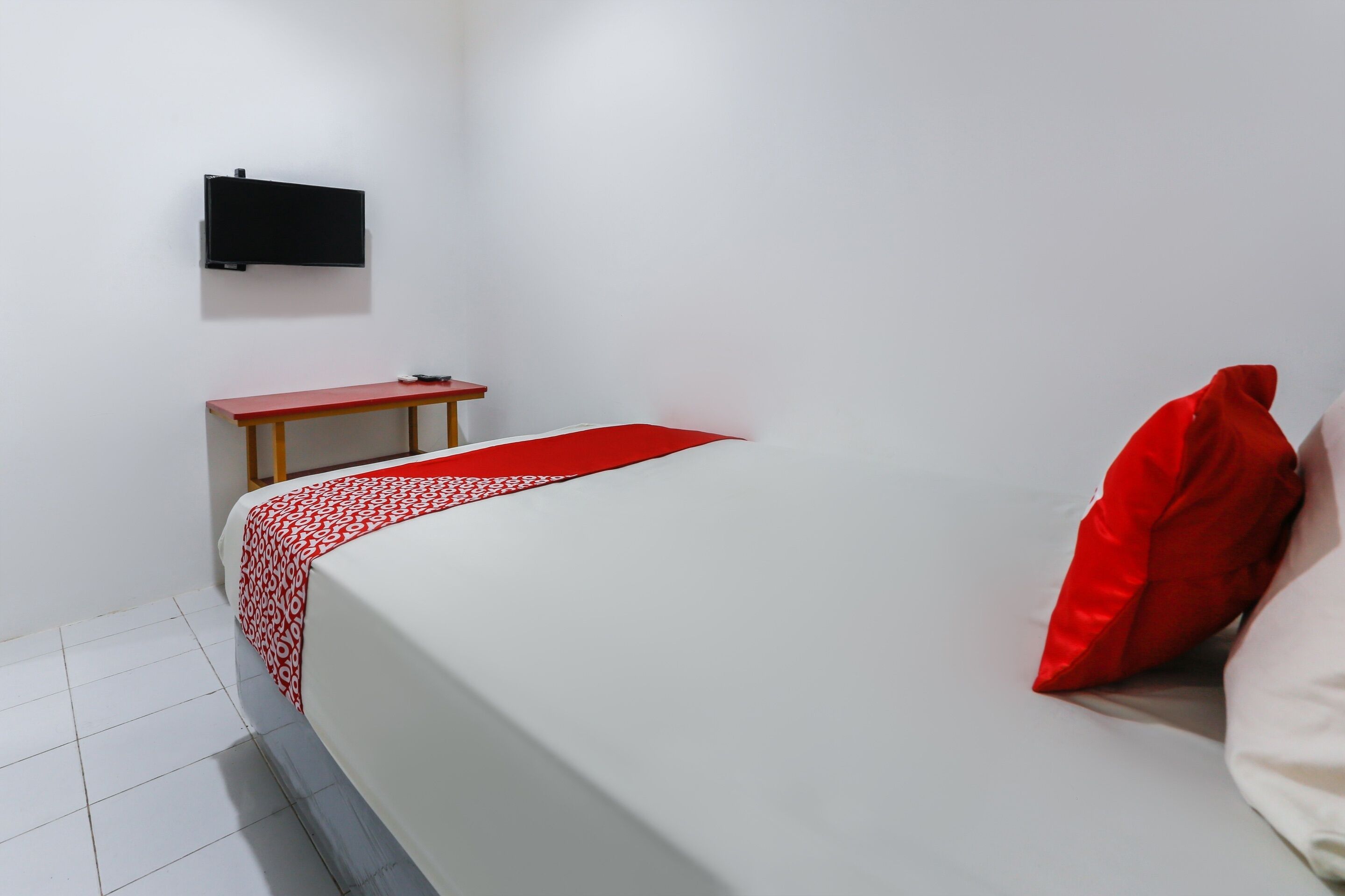 Habitación de hotel con cama, una mesita, televisor montado en la pared y una colcha con estampado rojo y blanco.
