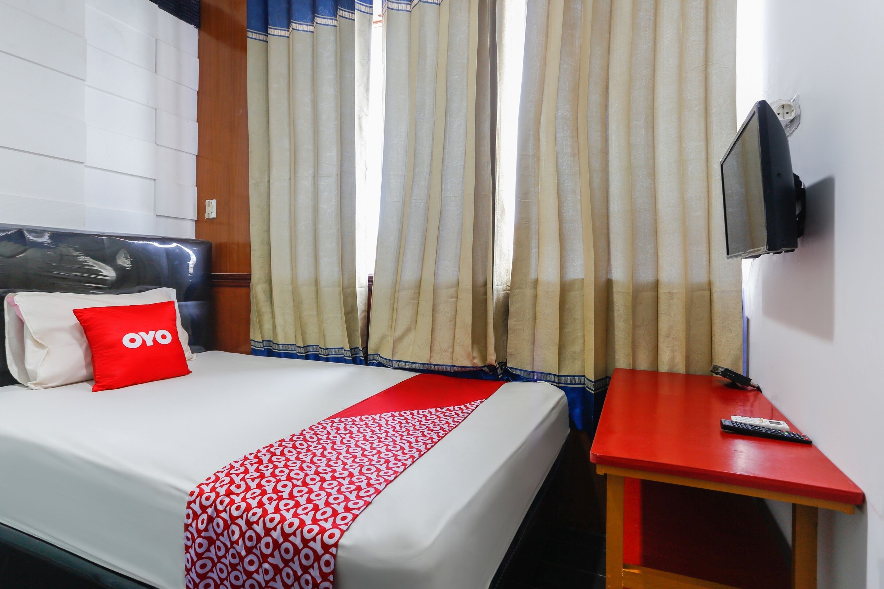 A hotel room with a bed, a red and white patterned bedspread, a red pillow with "OYO" text, a TV mounted on the wall, and a small wooden table with a remote control.