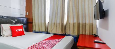 A hotel room with a bed, a red and white patterned bedspread, a red pillow with "OYO" text, a TV mounted on the wall, and a small wooden table with a remote control.