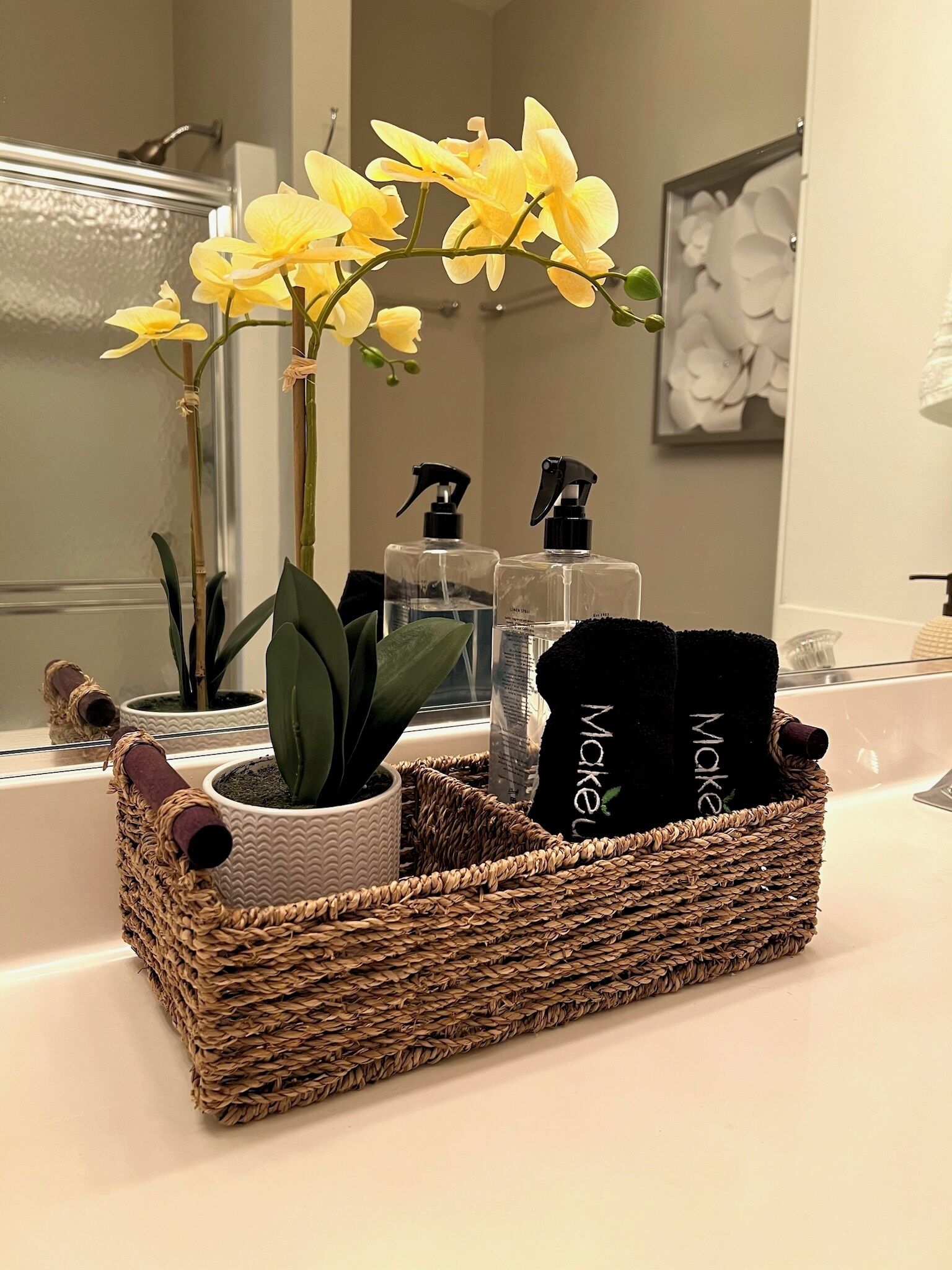 Um lavatório de casa de banho com uma cesta trançada contendo uma planta em vaso, uma toalha preta e um borrifador.