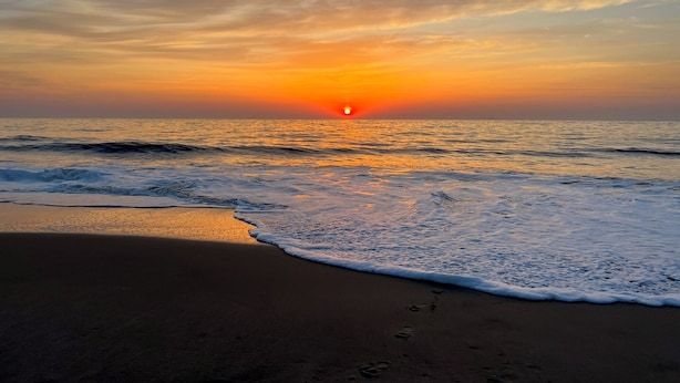 Ein Strand mit Sonnenuntergang über dem Meer, sanfte Wellen umspülen das Ufer.