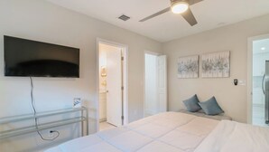 Une chambre à coucher avec un lit, un téléviseur fixé au mur, un ventilateur de plafond et une porte donnant sur une autre pièce.