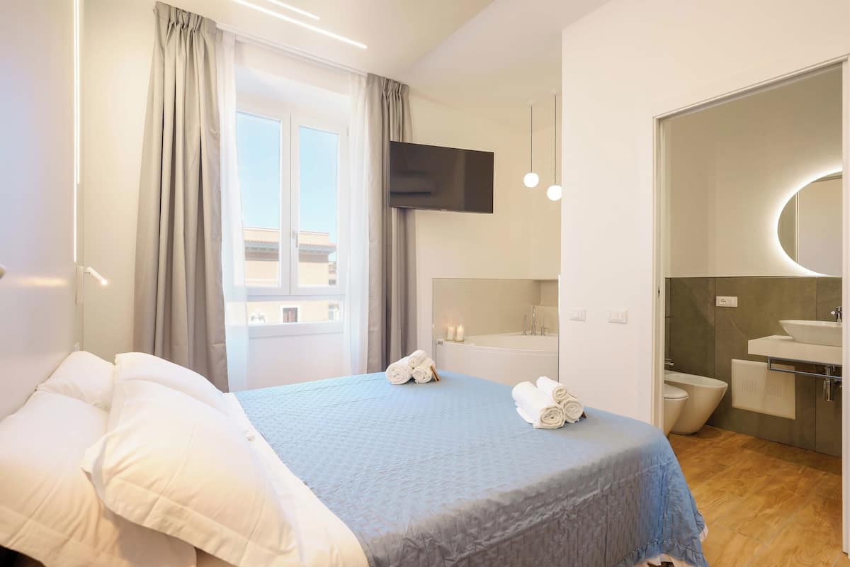 A modern hotel room with a large bed, a flat-screen TV, a bathroom with a modern sink and mirror, and a view of the city through the window.