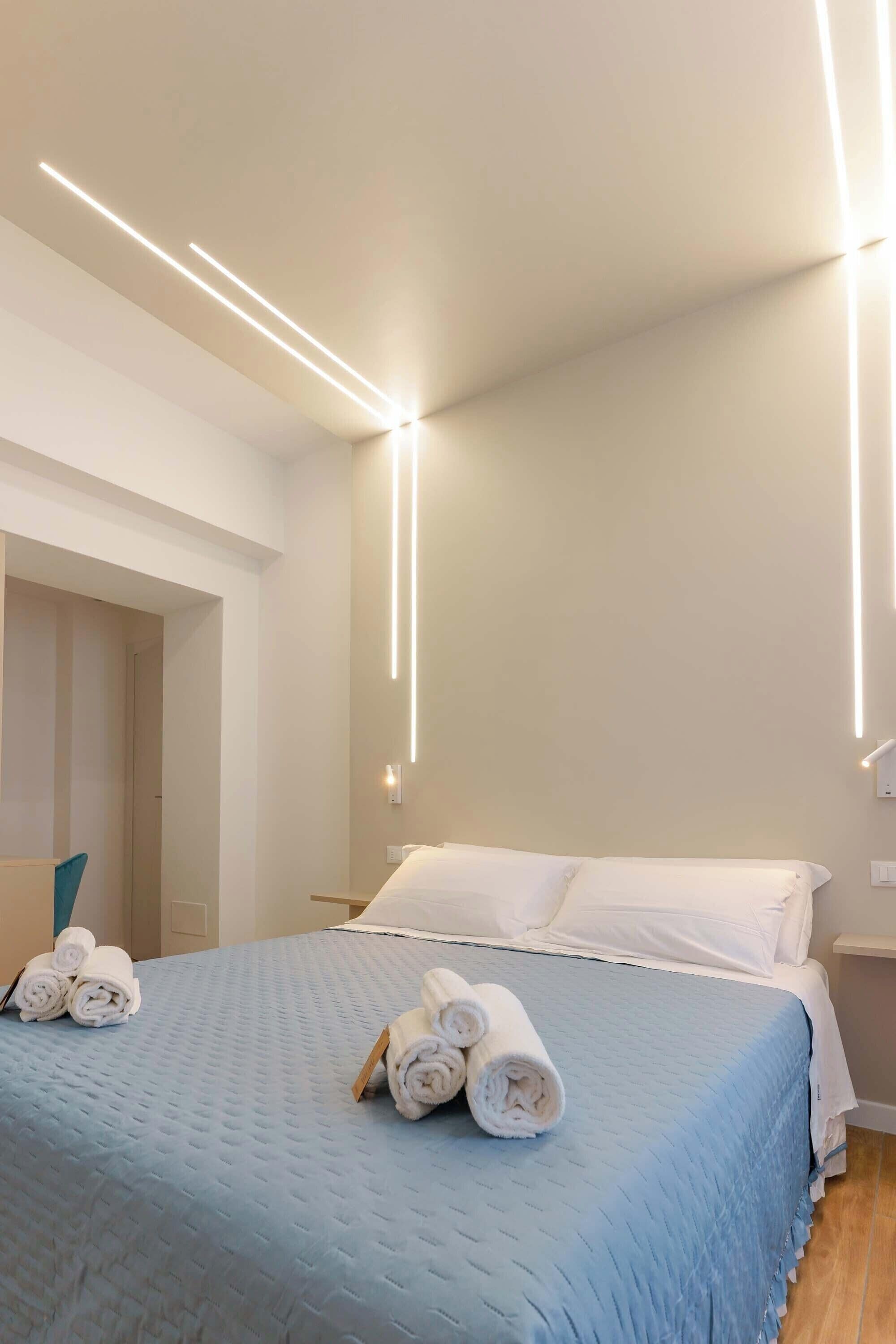 A modern hotel room with a neatly made bed, a blue quilted bedspread, and rolled towels on the bed.