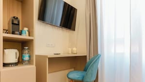Una habitación moderna con una televisión de pantalla plana en la pared, una mesa de centro de madera, una silla azul y un estante con una tetera, una botella de agua y una taza de café.