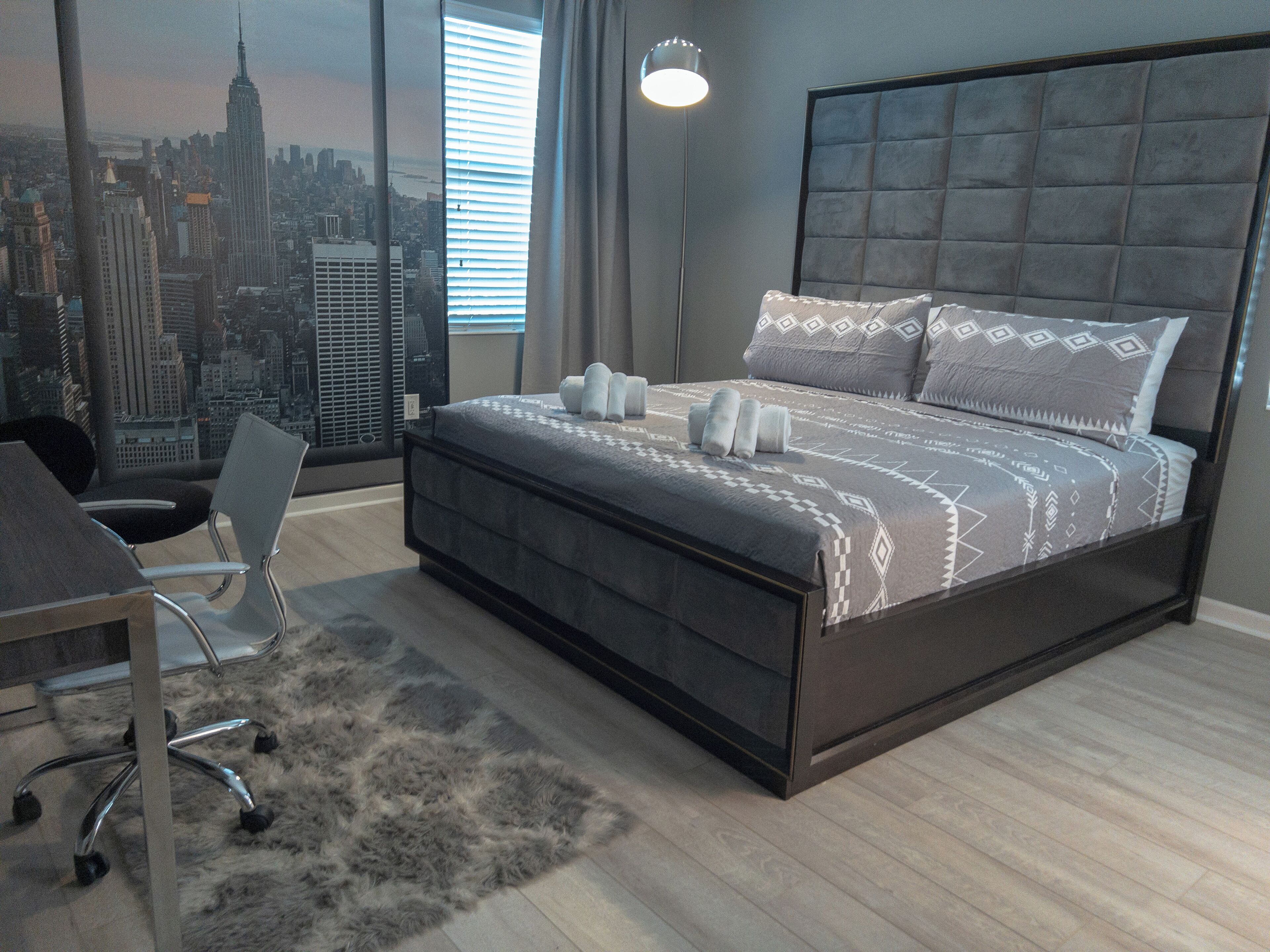 A modern bedroom with a cityscape mural, a large bed with patterned bedding, a desk, and a chair.