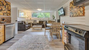 A modern kitchen with a built-in oven, a dining area with a wooden table and chairs, a sofa set, and a large window providing natural light.