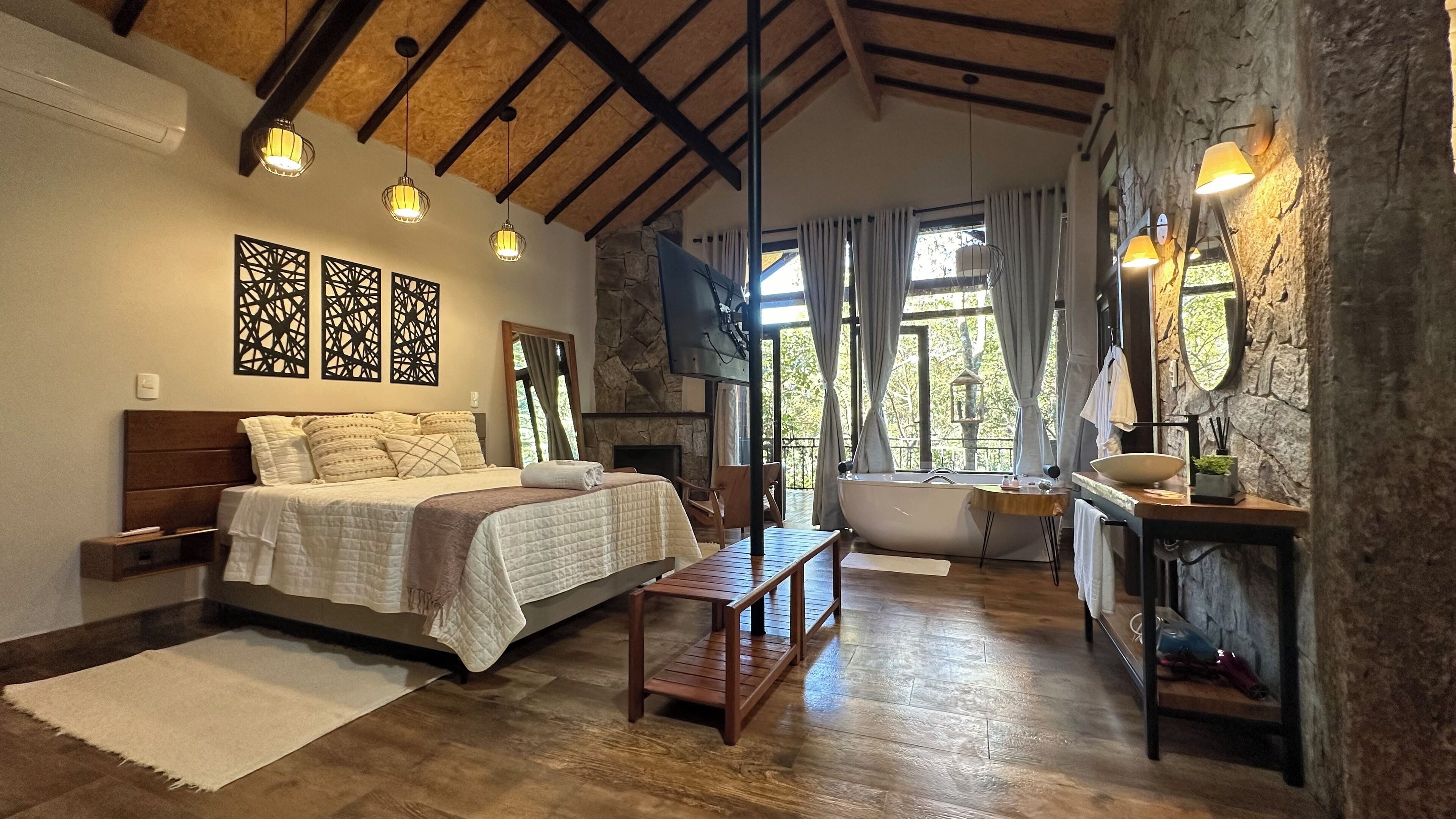 A bedroom with a bed, bedside tables, a wooden bench, a bathtub, a stone wall, and a large window with curtains.