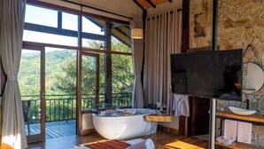 A bedroom with a large bed, a bathtub, a television, and a stone wall. - Monte Mauá Chalés (Resende)