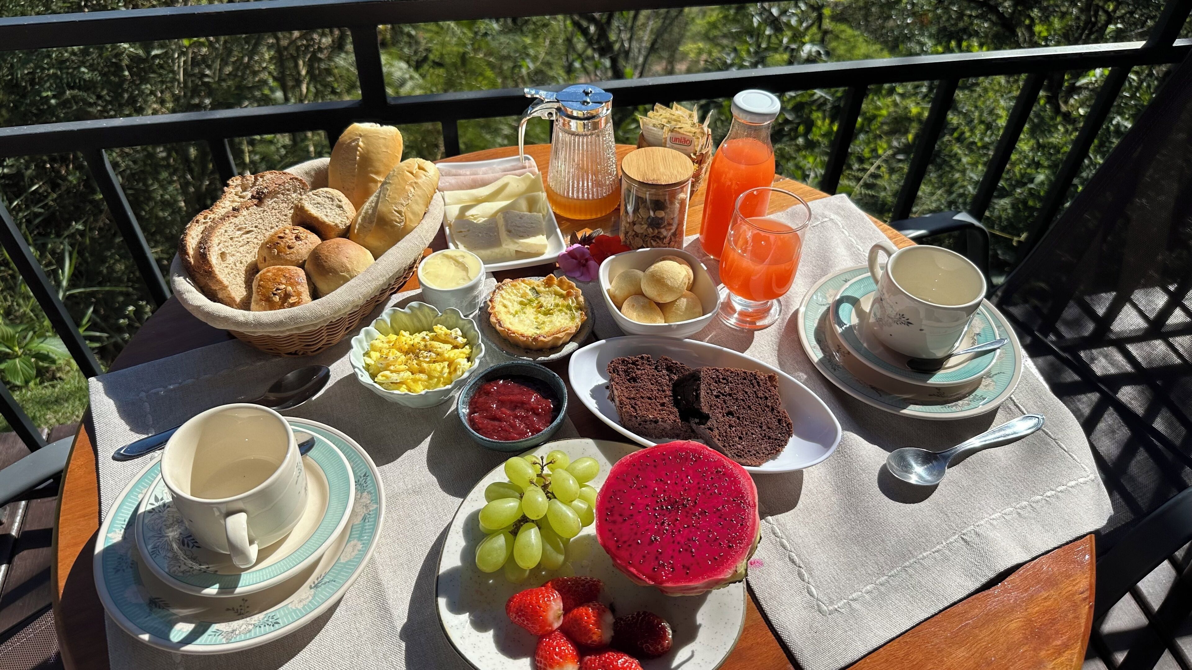 Una mesa de desayuno con diversos alimentos y bebidas, como pan, huevos, fruta y café, en un balcón con barandilla.