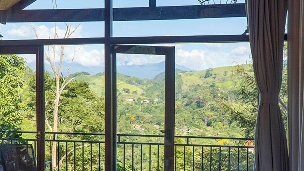 A room with a large window, a hot tub, and a wooden table with a bottle and glasses. - Monte Mauá Chalés (Resende)