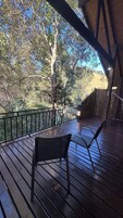 Una terraza de madera con dos sillas de metal, barandilla de madera y vista a árboles y colinas.
